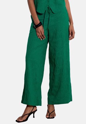 Woman wearing loose green pants with floral embroidery and black heeled sandals, with a black wristband on her left wrist.
