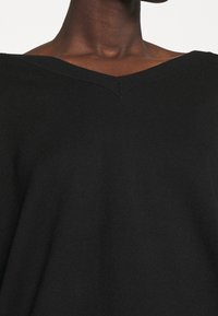 Svart stickad tröja med V-ringning, slät textur och subtila sömnadsdetaljer längs halsringningen och fållen.