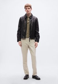 Μαύρο δερμάτινο μπουφάν με γιακά, ελιά πράσινο πόλο μπλουζάκι από κάτω, μπεζ παντελόνια και μαύρα μοκασίνια, δημιουργώντας ένα casual look.