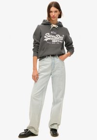 Felpa grigia con logo bianco, tasca a marsupio e cordino. I jeans a gamba larga di colore blu chiaro e le scarpe nere completano l'outfit.