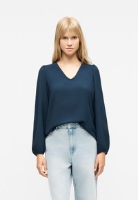 Νεαρή γυναίκα με μακριά ξανθά μαλλιά φοράει μπλε σκούρο μπλουζάκι V λαιμόκοψη με μακριά μανίκια και ανοιχτό μπ λε τζιν ψηλόμεσο, στέκεται μπροστά σε λευκό φόντο.