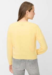 Gelber Strickpullover mit langen Ärmeln und rundem Ausschnitt, ausgestattet mit einer weichen Textur und einem körpernahen Saum. Wird mit grauer Jeans getragen.