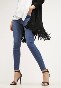 Jeans ajustados azules combinados con un cárdigan negro con flecos y tacones altos negros con tiras, mostrando una tela suave y un diseño ceñido.