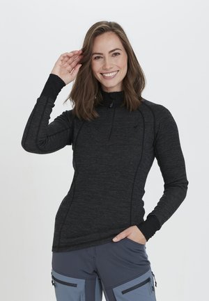 Zwarte longsleeve zip-up top met een hoge kraag en een getailleerd ontwerp. Kenmerkt zich door textuurstof en subtiele stikseldetails. Een model poseert met een glimlach.