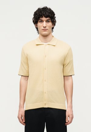 Mężczyzna o kręconych włosach, ubrany w beżową, dzianą koszulę z krótkim rękawem i guzikami oraz czarne spodnie, stojący na jednolitym białym tle.