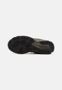 Semelle d'une chaussure de randonnée marron montrant une bande de roulement en caoutchouc noir avec des rainures profondes et des motifs de grip texturés sur un fond blanc.