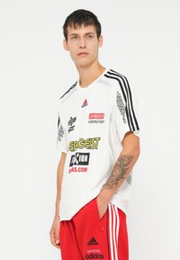 Bílý sportovní triko s černými a červenými akcenty, zdobené různými tištěnými logy. Krátké rukávy a kulatý výstřih.