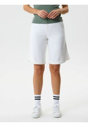 Persona vistiendo pantalones cortos blancos hasta la rodilla, zapatillas blancas, calcetas blancas con rayas negras y una camiseta verde, de pie contra un fondo liso.