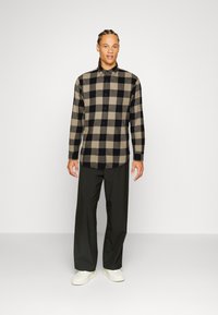 Camisa de franela a cuadros en beige y negro con mangas largas y cierre de botones, combinada con pantalones anchos oscuros y zapatillas blancas.