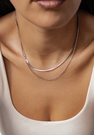 Silberne, doppellagige Halskette mit einer glatten, geschwungenen Kette und einer Perlenkette, die auf nackter Haut ruht und ihren metallischen Glanz hervorhebt.
