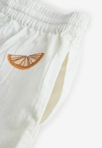 Valkoiset pellavashortsit, joissa on joustava vyötärönauha ja sivutaskut. Koristeena kirjailtu appelsiiniviipale. Pehmeä koostumus ja kevyt malli.