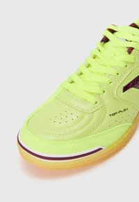 Neon dzeltenas sporta apavi ar ādas augšdaļu, perforētu purngalu un purpura akcentiem. Iekļauj izteiksmīgu zīmolu un caurspīdīgu gumijas zoli.