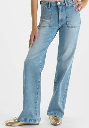 Jean large jambe couleur bleu clair avec grandes poches plaquées frontales, porté avec des ballerines dorées, visible de la taille aux pieds.