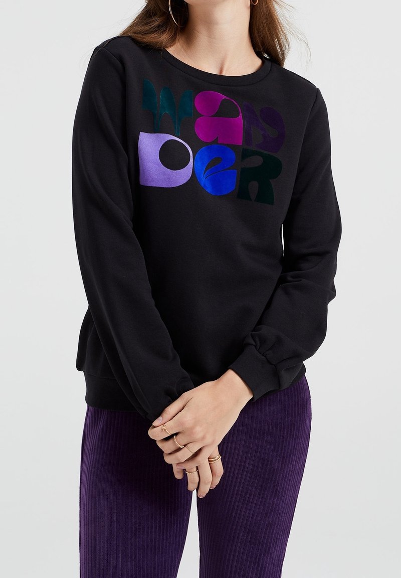 Femme portant un chemisier noir à manches longues avec des lettres abstraites colorées et un pantalon violet côtelé, les mains jointes devant, sur un fond clair uni.