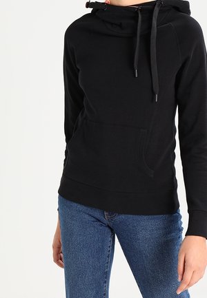 Osoba ubrana w czarną bluzę z kapturem i kieszenią z przodu oraz niebieskie dżinsy, pokazana od szyi do bioder na białym tle.