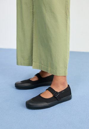 Pieds portant des baskets noires style Mary Jane avec une bride à boucle, associées à un pantalon large vert clair sur un tapis bleu.