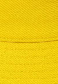 Geel emmerhoedje gemaakt van gestructureerde stof met een geribd patroon. Heeft duidelijke horizontale naden en een zachte, flexibele rand.