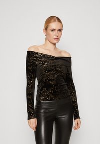 Top nero a spalle scoperte realizzato in morbido velluto, con motivi di stelle dorate, maniche lunghe e una vestibilità aderente. Indossato con leggings neri.