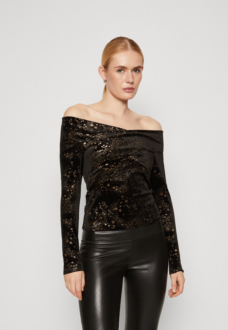 Top nero a spalle scoperte realizzato in morbido velluto, con motivi di stelle dorate, maniche lunghe e una vestibilità aderente. Indossato con leggings neri.
