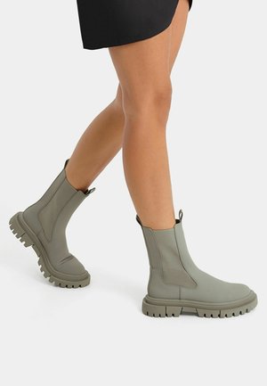 Des jambes portant des bottes à élastique gris clair, à semelle épaisse, arrivant mi-mollet, assorties à une jupe noire sur un fond clair.