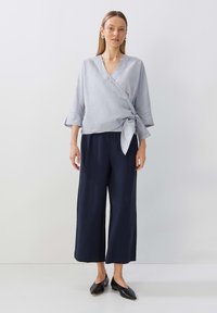 Top avvolgente grigio chiaro con maniche a tre quarti, legato in vita, abbinato a pantaloni a gamba larga blu navy e scarpe nere senza lacci.