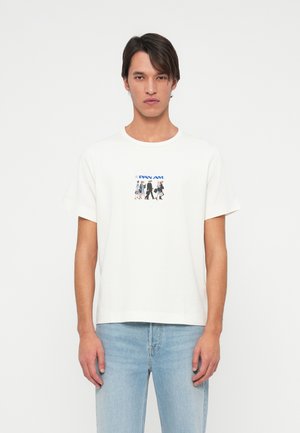 Mężczyzna w białym t-shircie z logo Pan Am i graficznym motywem w stylu vintage przedstawiającym stewardessy, połączony z jasnoniebieskimi dżinsami.