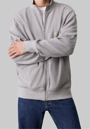 Sudadera con cremallera gris claro, de tejido texturizado, con puños y cuello acanalados, que presenta un pequeño logo en el lado izquierdo. Combinada con jeans oscuros.