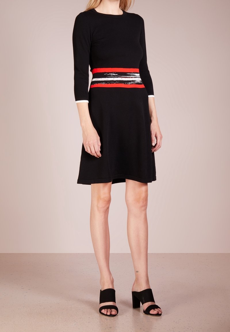 Femme portant une robe noire longueur genoux avec des manches trois-quarts et un détail de ceinture rayée rouge, blanche et noire, associée à des escarpins noirs ouverts à l'avant.