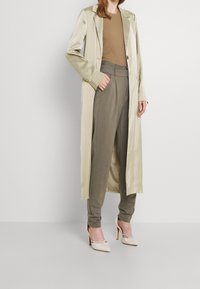 Beige satin long coat, brown fitted top, olive green tailored pants, and beige heels with a cutout design.