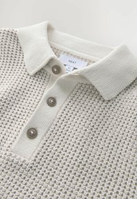 Cămașă polo cu guler, de culoare bej deschis, cu un model texturat, având trei butoane maro și o etichetă care afișează detalii despre marcă și mărime.
