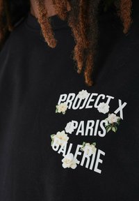 Zwarte t-shirt met een grafisch ontwerp waarop de tekst "PROJECT X PARIS GALERIE" staat, omringd door witte florale elementen.
