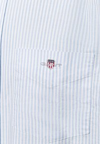 Lichtblauw en wit gestreept shirt met een borstzak en een geborduurde GANT-logo schild. De stof voelt glad aan.