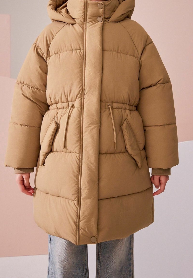 Enfant portant un manteau d'hiver rembourré beige avec une capuche, des boutons à l'avant, une taille à cordon et de grandes poches avant, assorti à un jean bleu.