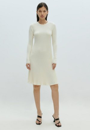 Geribbelde, langslijf jurk in crème, met een aansluitende bovenkant die uitloopt in een rok. Gecombineerd met zwarte sandalen met strappy. Gladde textuur.