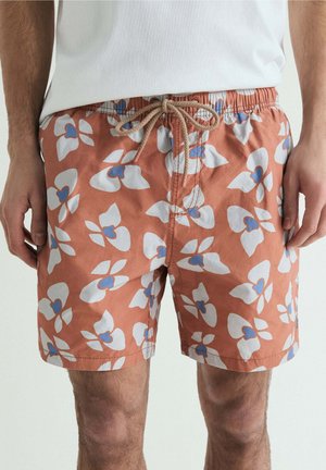 Bañador corto coral para hombre con estampado floral azul y blanco y cordón beige, combinado con camiseta blanca.