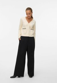 Kremowy kardigan z głębokim dekoltem w kształcie litery V, dwie przednie kieszenie z czarnymi akcentami, zestawiony z szerokimi czarnymi spodniami o gładkiej fakturze i dopasowanym kroju.