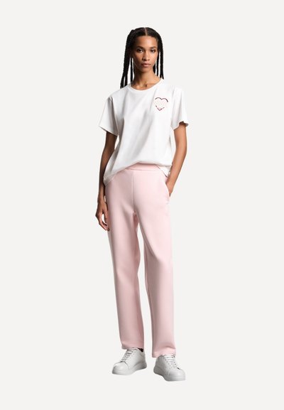 Maglietta bianca di cotone a maniche corte con grafica a forma di cuore, abbinata a pantaloni rosa chiaro in tessuto morbido e sneakers bianche.