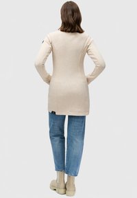 Béžová tunika s dlhým rukávom a priliehavým strihom, jemnou textúrou a bočnými rozparokmi; oblečená cez svetlo modré džínsy a krémové členkové čižmy.