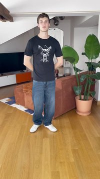 Svart T-skjorte med ørn-grafikk, blå jeans og hvite sneakers. Står på tregulv ved siden av en potteplante og en oransje sofa.