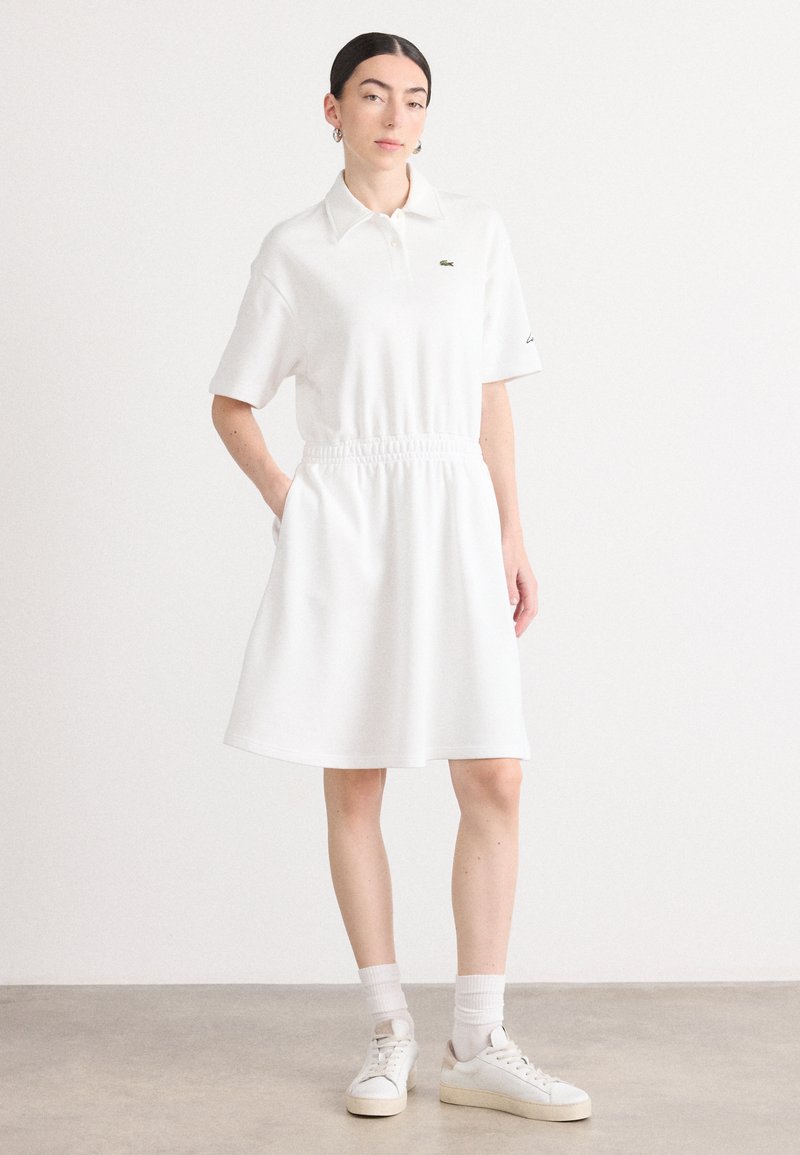 Balta polo suknelė su trumpomis rankovėmis, elastiniu juosmeniu ir A formos siluetu. Viršutiniame kairiajame krūtinės kampe yra žalias Lacoste logotipas.