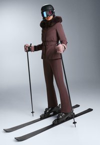 Skijaška odjeća u dubokoj bordo boji s kapuljačom obrubljenom krznom, uski kroj, gladak materijal, uparena s crnim skijama i čizmama, štapovi za skijanje u rukama.