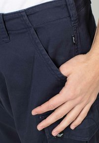 Pantalones cargo navy confeccionados en una mezcla de algodón, con bolsillos laterales y una pequeña etiqueta. Textura suave y ajuste entallado acentuado por costuras visibles.