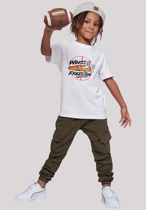 Weißes T-Shirt mit Grafik "Wings of Freedom" in Orange und Schwarz. Olivgrüne Cargo-Shorts. Weiße Sneaker mit strukturierten Akzenten. Kind, das einen Fußball hält.