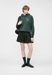 Sweat à capuche vert avec logo devant, jupe plissée noire, chaussettes vertes et chaussures noires. Design simple, coupe confortable et style décontracté.