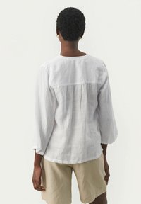 Personne avec des cheveux courts et bouclés, portant de grandes boucles d'oreilles en or, un blouse blanche à manches longues ample et un short beige, se tenant de dos.