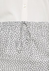 Biała koszula na guziki z fakturowanej tkaniny, z paskiem ściągającym w talii, zdobiona szarym wzorem kwiatowym na jasnym tle.