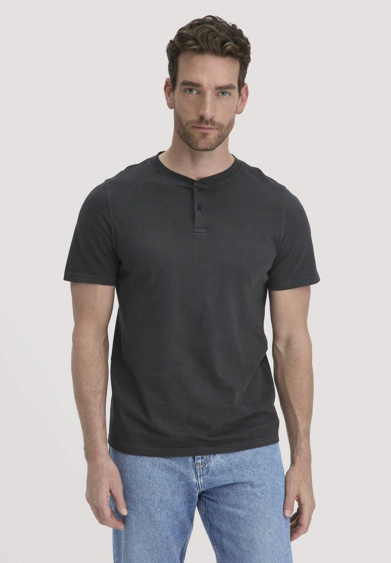 Mann mit kurzen lockigen Haaren, der ein schwarzes kurzärmliges Henley-Shirt und hellblaue Jeans trägt, steht vor einem einfarbigen hellen Hintergrund.