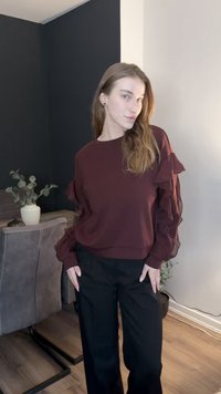 Μπορντό φούτερ με μουσελίνες μανίκια με βολάν, σε συνδυασμό με μαύρο παντελόνι φαρδύ. Μινιμαλιστική διακόσμηση και γλάστρα στο φόντο.