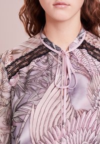 Mujer vistiendo una blusa floral semitransparente con borde de encaje negro y una cinta rosa anudada en el cuello, mostrando el cuello y la parte superior del pecho.