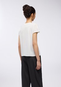 T-shirt bianca in cotone con scollatura rotonda e maniche corte, abbinata a pantaloni neri larghi, vista da dietro su uno sfondo neutro.